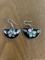 Photo of a pair of semi-circular earrings with iridescent mother of pearl inlay arranged in floral shapes on a black base, shown flat on a wood surface.