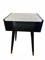 Front angled view of the black nightstand with faux marble top showing the open storage compartment and splayed tapered legs with brass caps.