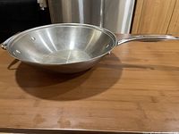 Side angle image of the stainless steel wok showing the long handle and small loop handle on wooden table.