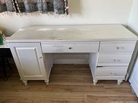 Front view of a white wooden desk showing three drawers on the right, one central drawer, and cabinet on the left, standing on a wooden floor.