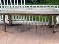 Full view of the large handmade wooden work table showing its length and sturdy legs on a concrete surface with white fence background.