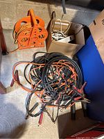 Photo showing extension cord reel, box with white cords, and multiple jumper cables with clamps on the floor