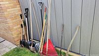Photo showing multiple garden tools leaned against outdoor wall including 5 winter shovels, red plastic fan rake, metal hard rake, axe, and shovels.