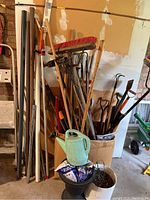 Photo showing assorted garden tools in a bucket including brooms, rakes, hoes, shovels, metal poles, plastic plant pots and a green watering can.