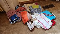 Lot of vintage ladies aprons folded on carpet floor showing various colors and patterns including blue floral, red checkered, white with pink design, orange and others.