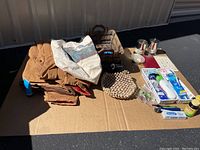 Overview of lot showing brown jacket, woven pad, wooden bin, silver candle holders, framed certificate, and personal care items arranged on cardboard.