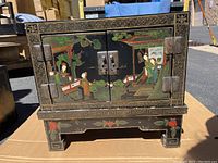 Front view of the black lacquer chest showing two doors with painted scene of figures outdoors with plants, bordered by geometric patterns in gold.