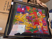 Framed colorful abstract painting featuring vibrant shapes and fruit, under glass, with black and gold frame.