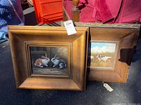 Two framed oil paintings placed on ground; one depicting two rabbits facing each other, the other showing birds in nature. Both frames are gold-tone with crackle finish.