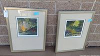 Two framed watercolor paintings side by side against a brick wall showing nature scenes.