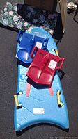 Photo of blue foam snow boogie board with yellow handles and two plastic sleds, one blue and one red, positioned on it.
