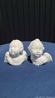 Front view of both white porcelain cherub angels displayed side-by-side on blue surface against dark background, showing facial detail and winged shoulders.