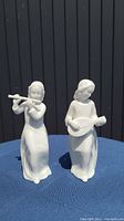 Two white porcelain figurines on a blue surface against dark slatted background; one playing flute, the other playing lute; both approx 5.5 inches tall, one broken.