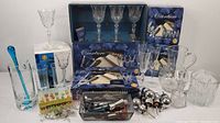 Barware collection including boxed Overture Capri Crystal champagne flutes and large goblets, glass pitchers with blue glass stirrers, ice bucket, metal wine stoppers, and colorful cocktail markers.