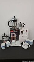 Overview of coffee set including two French presses, stovetop espresso maker, ceramic teapot, mugs, and saucers.