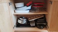 Cabinet shelf with stacked white Corning Ware dishes, Pyrex glass dishes with red lids, clear glass rectangular baking dish on lower shelf. Metal bakeware including muffin pan, loaf pans, cookie sheets and other baking trays are on lower shelf.