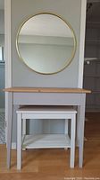 Gray table with wood top, smaller white table underneath, round gold-framed mirror above on wall
