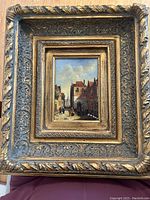Frontal view of framed painting showing ornate gilt frame and signed village scene artwork