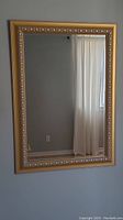 Full view of the rectangular gold coloured framed mirror mounted on a wall showing clear reflective surface with a curtain reflected.