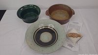 Lot of pottery items including three bowls in sage, dark green, and brown colors and a clay fish bun warmer with info sheet on a white cloth.
