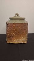 Front view of large handmade ceramic pottery jar with lid showing decorative carved floral designs and loop handle on lid