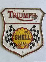 Front view of shield-shaped cast iron advertising sign with Triumph and Shell logos and racing flags.
