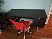 Photo of a large black desk with a matte finish on a wooden floor next to a pale blue wall. A red-orange swivel chair with metal armrests and base is placed in front of it.