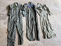 Wide view of all three flight suits laid flat on a concrete floor showing overall condition and color variation between olive green and tan suits.