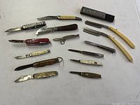 Overview of 12 vintage pocket knives/tools and 2 straight razors with original box, all laid out on white surface.