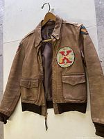 Front view of brown leather A-2 jacket on hanger showing two front flap pockets, zipper closure, ribbed hem and cuffs, round left-chest patch and shield right-shoulder patch.