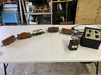 Side view of six model train cars and transformer controller laid out on a white table in a garage setting.