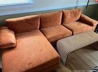 Orange retro fabric couch with chaise lounge, three back cushions, and wide armrest next to wicker coffee table on hardwood floor under window