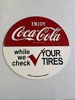 Front view of round porcelain Coca-Cola sign with red and white background, advertising Coca-Cola and tire checking service.