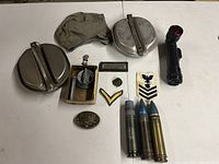 Photo showing all lot contents: two aluminum mess kits, three 30MM shell casings, several rank patches, a flashlight, a belt buckle, a small insignia, and fabric pouch.