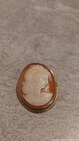 Front view of the oval cameo brooch showing the carved white female profile on a brown background surrounded by a gold-tone frame.