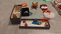 Wide view of assorted Christmas pins, earrings, and button in trays with visible variety of designs including bells, trees, and holiday motifs.