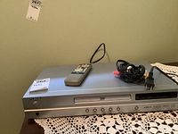 Koss DVD player shown with remote control on top, power cable coiled beside it, placed on a lace tablecloth.