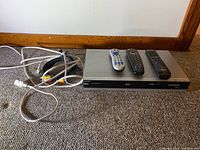 Philips DVD player/recorder with three remote controls and cables on carpeted floor against a wall.