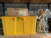 Yellow plastic storage bin and cardboard box visible, with resin statue figures behind them on a shelf in a garage with unfinished walls.