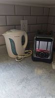 Photo showing Proctor Silex white electric kettle placed beside Hamilton Beach stainless steel 2-slice toaster on a kitchen countertop with a tiled backsplash.