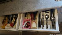Top opened drawers showing assorted cutlery and kitchen utensils including spoons, forks, knives with flower design handles, can opener, ladles, tongs, grater, and sharpener.