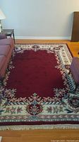 Full view of rectangular deep red wool area rug with floral border and fringe on hardwood floor beside furniture.