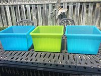 Three square plastic storage bins placed on an outdoor metal table. Two bins are light blue and one is green, showing their general appearance and color.