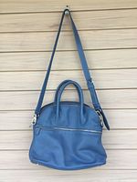 Front view of blue leather purse with detachable strap and two handles hanging on a hook against a wood wall.