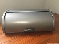 Front view of a matte grey metal Brabantia bread box with roll-top lid on a wooden surface