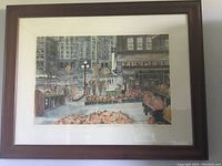 Framed print showing a detailed market scene with vendor baskets and buildings, in a dark wooden frame with white matting.