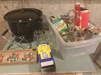 Photo showing a large black enamel canning pot with lid, multiple clear glass canning jars, assorted metal lids and rings in a plastic bin, jar lifter tool, and boxes with canning supplies.