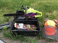 Chainsaw, open black plastic carrying case showing spare bar, chains, sharpening file, and safety helmet on grass