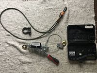 Manual come-a-long hand winch, steel cable with hooks and additional small shackle, shown next to black plastic carrying case designed to hold the tool and accessories.