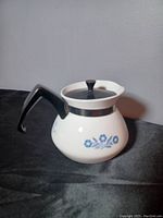 Side view of the white ceramic teapot with blue cornflower floral pattern, black handle and metal band near spout area, placed on black cloth background.